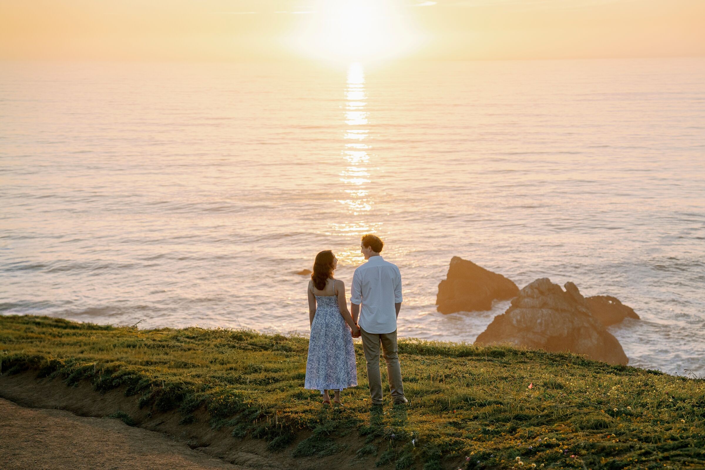 Melissa and Kye holding hands at a cliff edge, looking out at the sun over the ocean