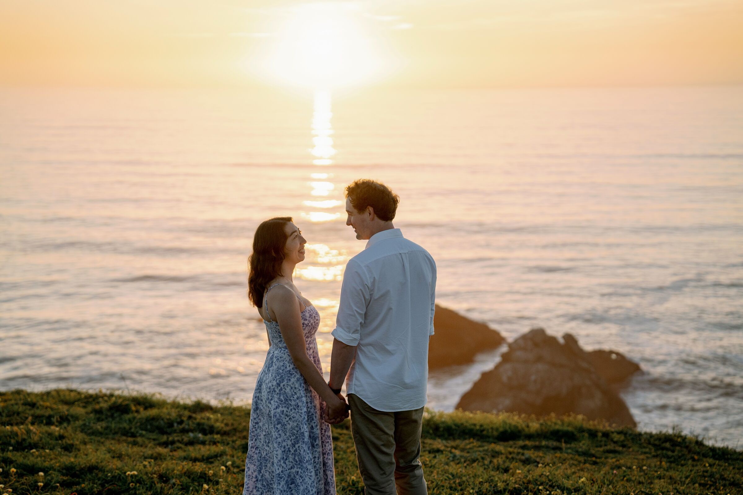 Melissa and Kye facing each other on a clifftop, the sun setting over the ocean behind them